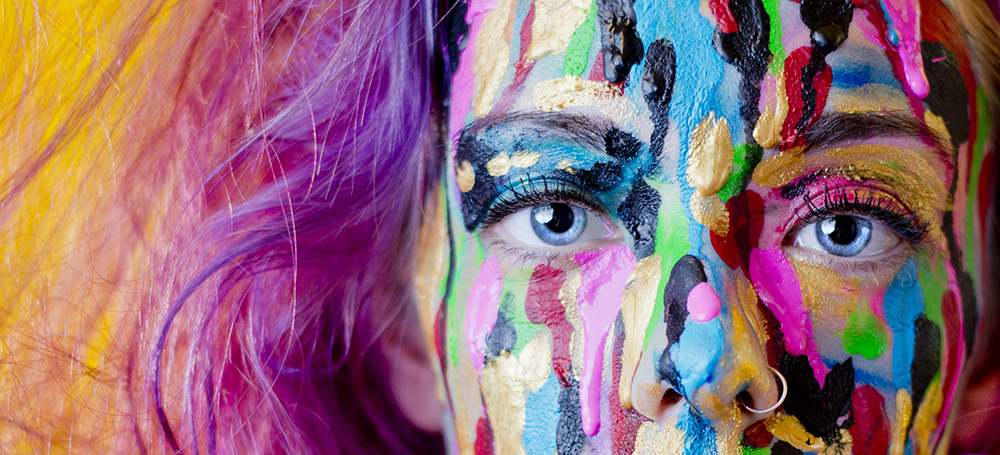 Ein Portrait einer Frau mit bunt bemaltem Gesicht und leuchtend blauen Augen.